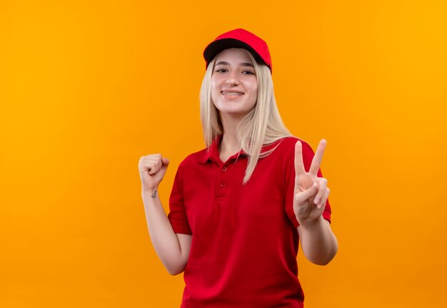 Sourire de livraison jeune fille portant un t-shirt rouge et une casquette en orthèse dentaire montrant la paix et oui des gestes sur fond orange isolé