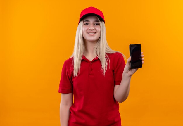 Sourire de livraison jeune fille portant un t-shirt rouge et une casquette en orthèse dentaire montrant le téléphone à la caméra sur fond orange isolé