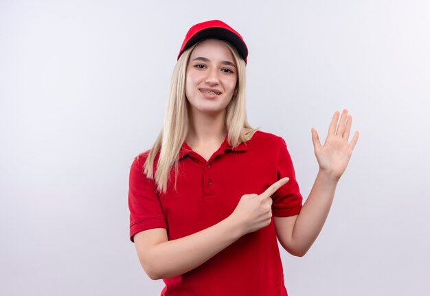 Sourire de livraison jeune fille portant un t-shirt rouge et une casquette en orthèse dentaire points à portée de main sur fond blanc isolé
