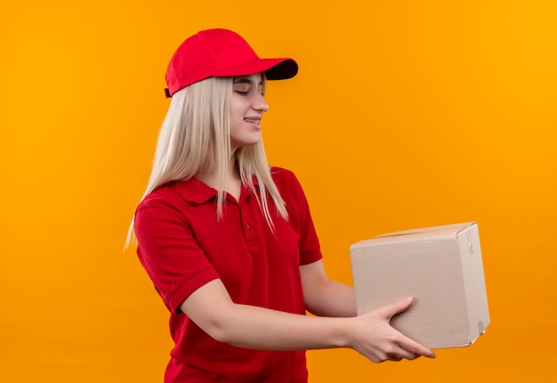 Sourire de livraison jeune fille portant un t-shirt rouge et une casquette en orthèse dentaire tenant la boîte à côté sur fond orange isolé