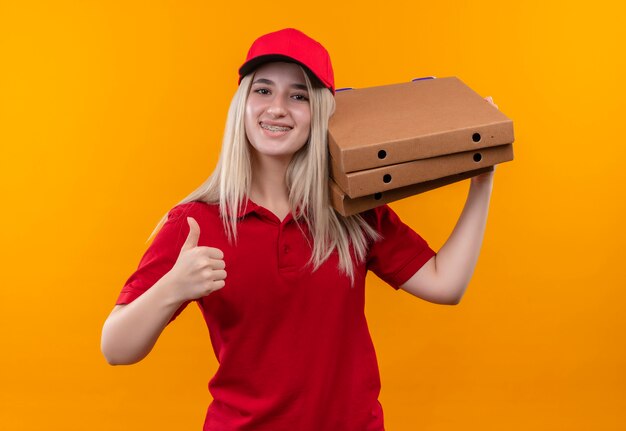 Sourire de livraison jeune fille portant un t-shirt rouge et une casquette en orthèse dentaire tenant une boîte à pizza sur l'épaule son pouce vers le haut sur fond orange isolé