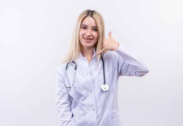 Sourire médecin jeune fille blonde portant un stéthoscope et une robe médicale en orthèse dentaire montrant le geste d'appel sur fond blanc isolé