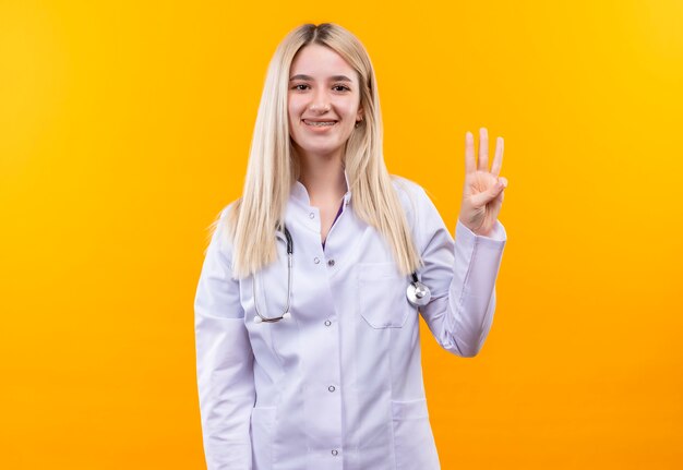 Sourire médecin jeune fille portant un stéthoscope en blouse médicale et orthèse dentaire montrant trois sur fond jaune isolé