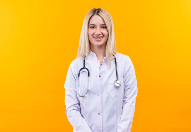 Sourire médecin jeune fille portant un stéthoscope en robe médicale et appareil dentaire sur fond jaune isolé