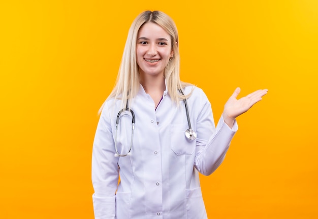 Sourire médecin jeune fille portant un stéthoscope en robe médicale et attelle dentaire pointe à côté sur fond jaune isolé