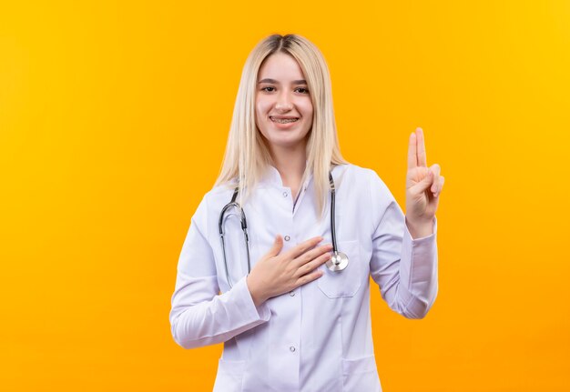 Sourire médecin jeune fille portant un stéthoscope en robe médicale et orthèse dentaire montrant deux sur fond jaune isolé