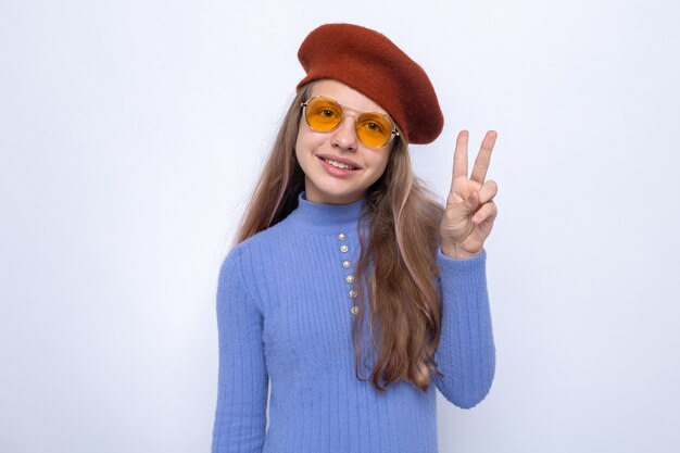 Sourire montrant un geste de paix belle petite fille portant des lunettes avec un chapeau isolé sur un mur blanc