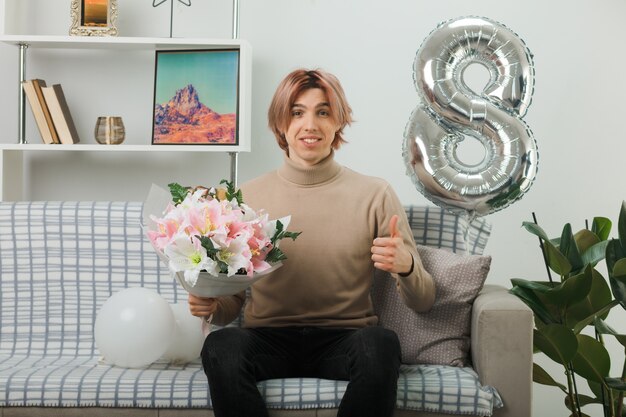 Sourire montrant le pouce vers le haut beau mec le jour de la femme heureuse tenant un bouquet assis sur un canapé dans le salon