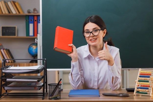 sourire montrant les pouces vers le haut jeune enseignante tenant un livre assis au bureau avec des outils scolaires en classe