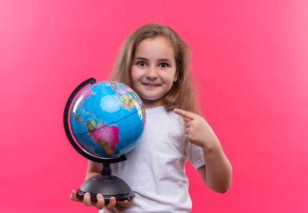 Sourire petite écolière portant un t-shirt blanc tenant un globe sur fond rose isolé