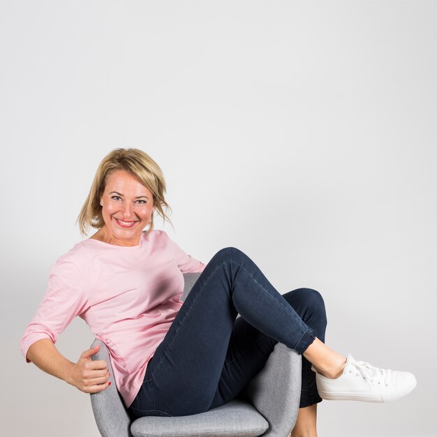 Sourire portrait d&#39;une femme mature assise sur un fauteuil gris sur fond blanc
