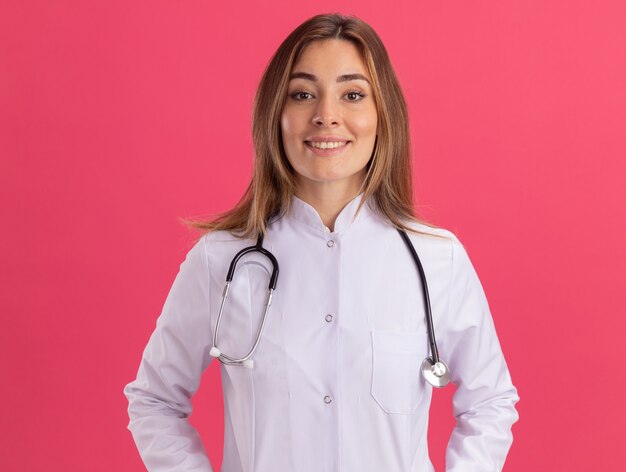 Sourire regardant devant jeune femme médecin portant une robe médicale avec stéthoscope isolé sur mur rose