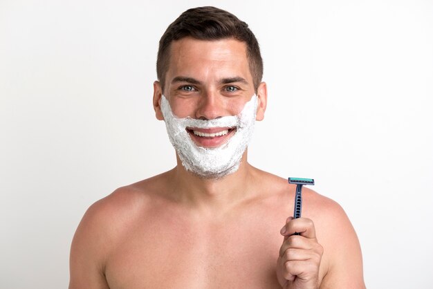 Sourire torse nu jeune homme avec mousse appliquée et tenant rasoir debout sur fond blanc