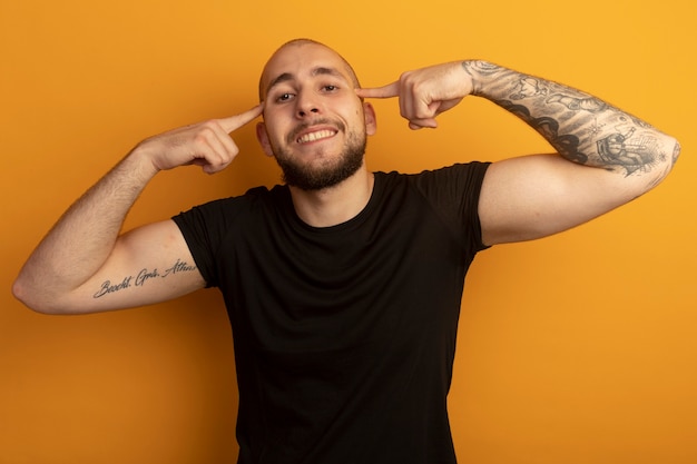 Photo gratuite sourire tout droit jeune beau mec portant une chemise noire mettant le doigt sur le temple isolé sur orange