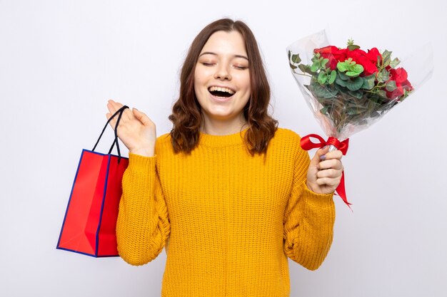 Sourire avec les yeux fermés belle jeune fille le jour de la femme heureuse tenant un sac-cadeau avec bouquet isolé sur mur blanc