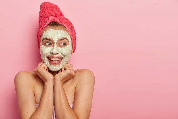 Spa girl étale une texture boueuse sur le visage, a l'air heureux, garde les mains sous le menton, détourne le regard avec le sourire, hydrate et calme la peau, porte une serviette rouge sur la tête, production excessive d'huile, se tient nue à l'intérieur