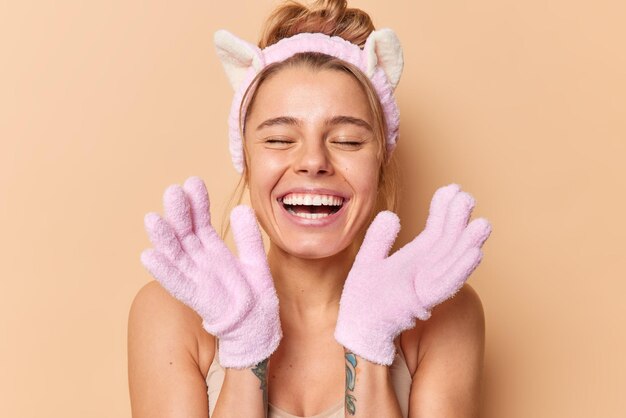 Spa de soins personnels et concept de douche. Une jeune femme positive sourit avec joie utilise des gants de bain pour laver et enlever les cellules mortes de la peau porte des poses de bandeau sur fond beige.