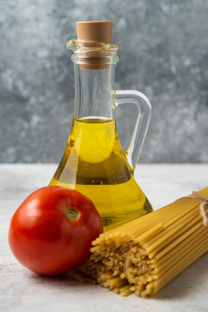Spaghetti sec, bouteille d'huile d'olive et tomate sur table blanche.