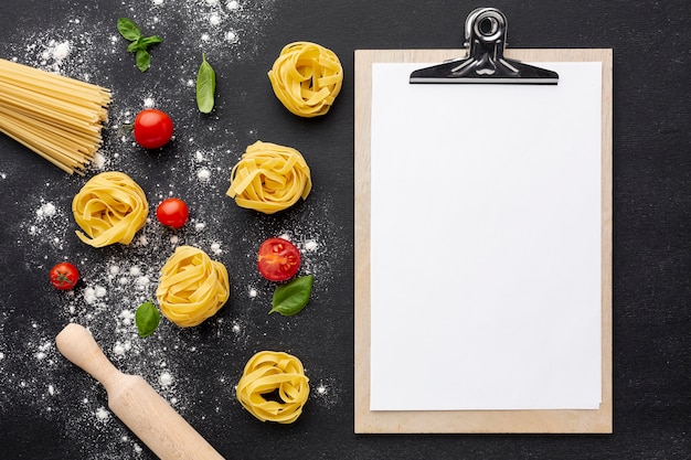 Spaghettis tagliatelles non cuites sur fond noir avec maquette de rouleau à pâtisserie et de presse-papiers aux tomates
