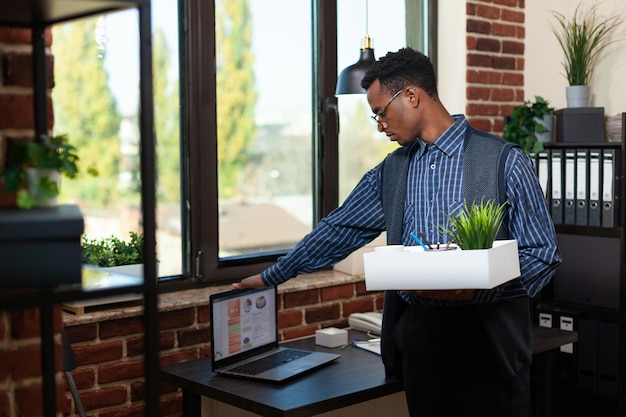 Un spécialiste du marketing licencié ferme un ordinateur portable avec des analyses commerciales tout en tenant un plateau avec des effets personnels avant de partir. Employé de bureau licencié arrêtant l'ordinateur portable après avoir ouvert le bureau.