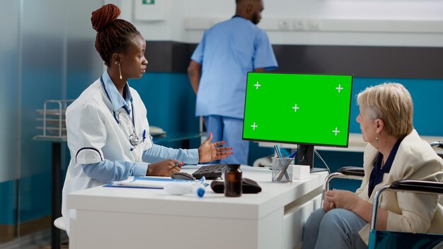Spécialiste pointant vers un ordinateur avec écran vert lors du contrôle avec une vieille femme en fauteuil roulant. Médecin et patient regardant l'arrière-plan de la clé chroma avec un fond de maquette isolé.