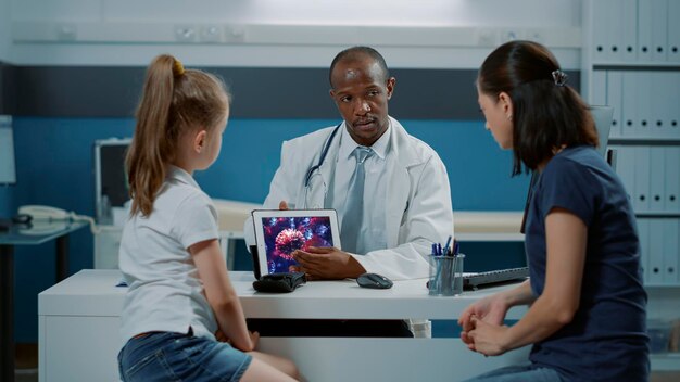 Spécialiste de la santé montrant une illustration de virus sur une tablette numérique à une femme et une fille. Pédiatre expliquant l'animation du coronavirus sur l'écran du gadget, donnant des conseils à la mère et à l'enfant au bureau.