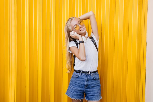 Spectaculaire femme mince tenant un casque blanc. Portrait en plein air de fille insouciante blonde posant sur fond jaune.