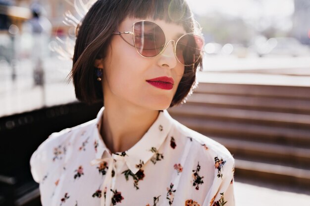 Spectaculaire jeune femme en blouse blanche posant dans la rue Incroyable fille brune à lunettes de soleil debout sur fond de ville