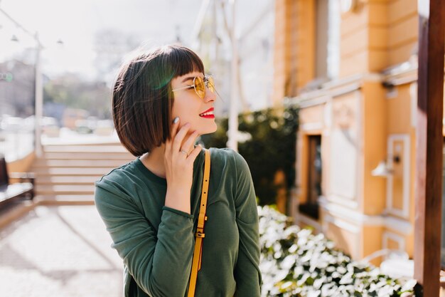 Spectaculaire jeune femme souriante en se promenant dans la ville par bonne journée chaude. Portrait en plein air du modèle féminin insouciant aux cheveux noirs raides.