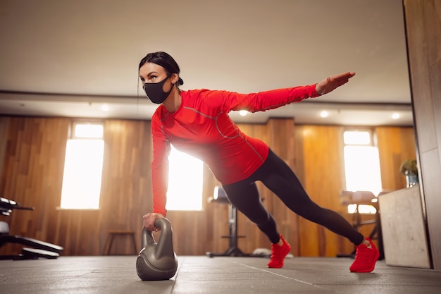Sport girl in mask de coronavirus faisant des planches kettlebells