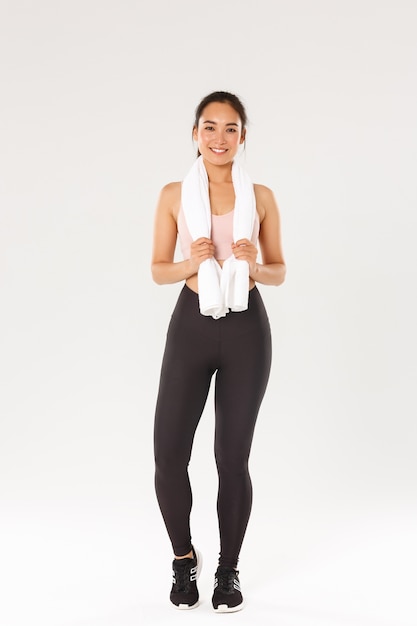 Sport, gym et concept de corps sain. Sur toute la longueur de la fille mince mignonne souriante, entraîneur de fitness ou sportive après des exercices dans la salle de gym, debout avec une serviette enroulée autour du cou, fond blanc.