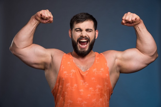 Photo gratuite sportif montrant ses muscles sur un mur bleu foncé.