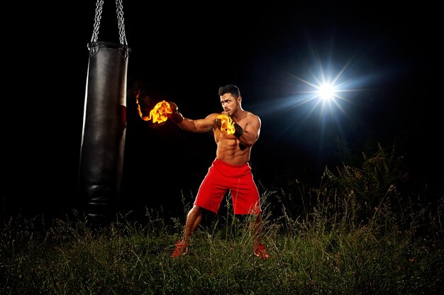 Sportif s'entraîne sur les gants de boxe corps musclé de fond de nuit noire dans le feu la formation de nuit d'entraînement en espace ouvert sur le coup de pied de boxe d'herbe du sac de boxe noir à droite