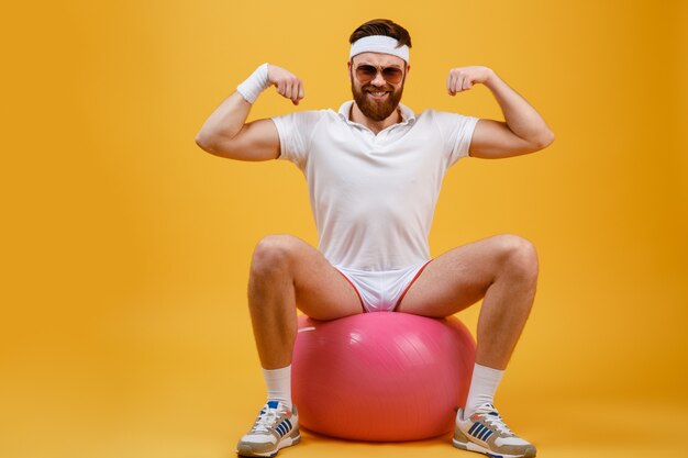 Sportif souriant assis sur un ballon de fitness et montrant les biceps