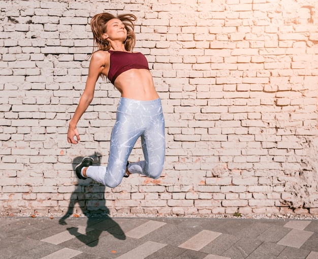 Photo gratuite sportive jeune femme sautant en l'air avec joie contre le mur de briques blanches