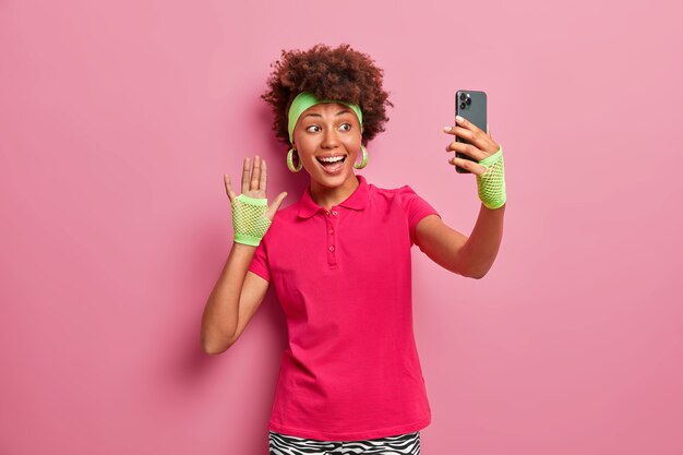 Une sportive positive en vêtements actifs fait des vagues à la caméra du smartphone, prend un selfie, envoie une photo aux abonnés, est de bonne humeur, fait un geste de salutation, sourit à l'écran mobile