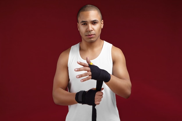 Sports, fitness, mode de vie sain et concept d'entraînement. Photo de jeune homme métis déterminé avec visage rasé en douceur et tête chauve mettant le bandage de boxe avant la formation avec sac de frappe