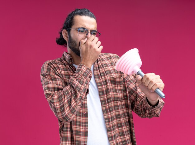Squeamish jeune beau mec de nettoyage portant un t-shirt tenant et regardant le nez de fermeture du piston isolé sur le mur rose