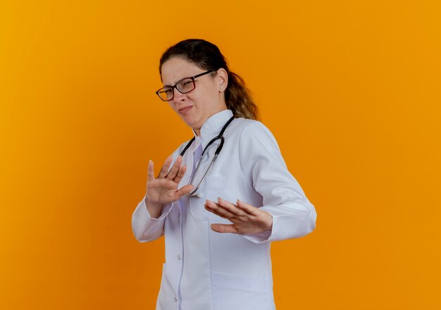 Squeamish jeune femme médecin portant une robe médicale et un stéthoscope avec des lunettes tenant les mains isolées
