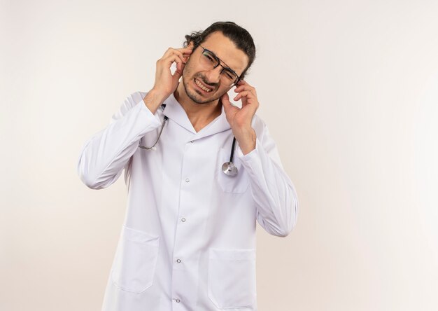 Squeamish jeune médecin de sexe masculin avec des lunettes optiques portant une robe blanche avec stéthoscope fermé les oreilles sur un mur blanc isolé avec copie espace