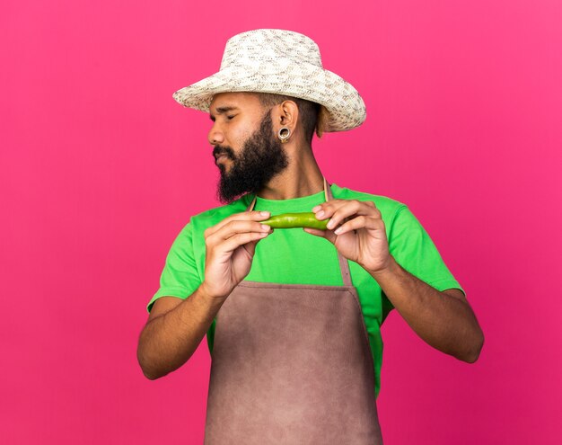 Squeamish avec les yeux fermés jeune jardinier afro-américain portant un chapeau de jardinage tenant du poivre