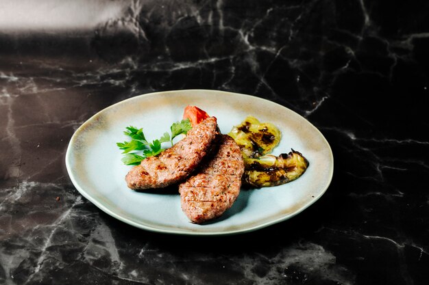 Steak de viande avec des aubergines grillées, du persil et des tomates dans une assiette blanche.