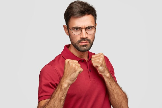Stern grave homme mal rasé garde les mains dans les poings, prêt à se battre avec le concurrent, regarde sous les sourcils, a déplu l'expression, vêtu d'un t-shirt rouge décontracté, se dresse contre un mur blanc