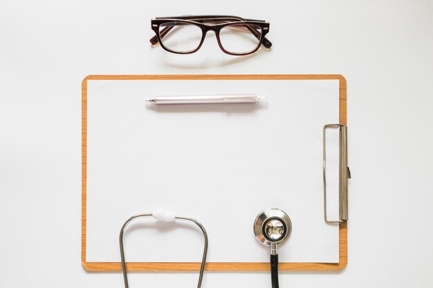 Stéthoscope; stylo et lunettes sur presse-papiers avec papier sur fond blanc