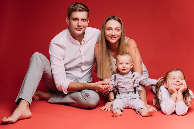 Stock photo de belle famille caucasienne complète avec mère et père assis sur le sol avec bébé fils et fille mignonne souriante allongée sur le sol. Découpe sur rouge.