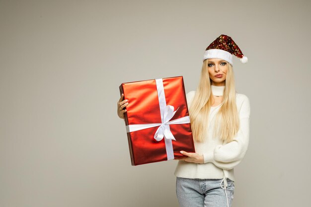 Stock photo de jolie fille blonde en bonnet de Noel, pull blanc et jeans bleu clair tenant un cadeau de Noël enveloppé et regardant la caméra avec une expression faciale douteuse.