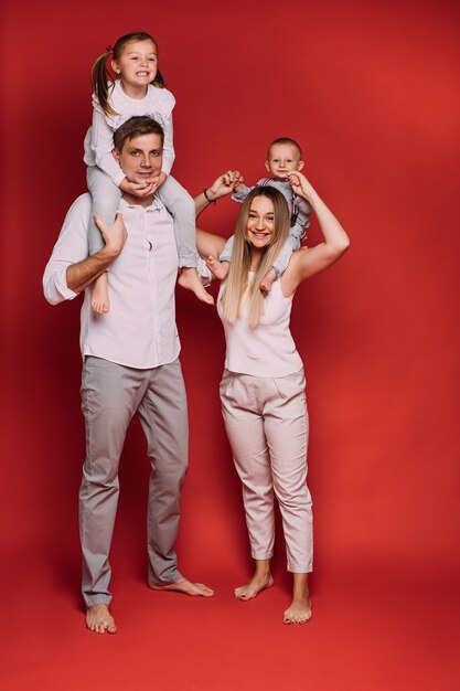 Stock photo de mère aimante avec bébé fils et papa avec fille assise sur les épaules et souriant joyeusement sur fond rouge.