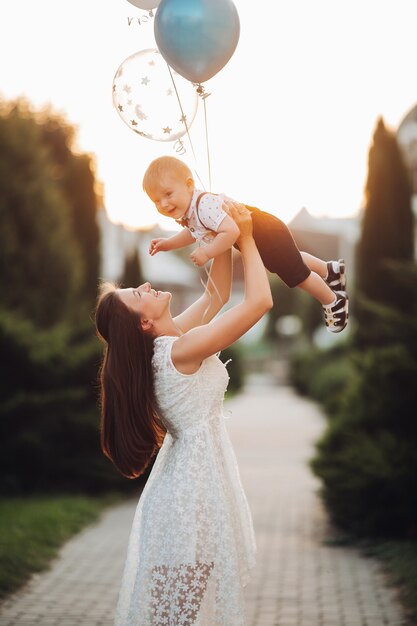 Stock photo d'une mère aimante dans une belle robe d'été blanche élevant son fils avec des ballons à air gonflables dans l'air dans un joli jardin en arrière-plan flou. Célébrer l'anniversaire de son fils à l'extérieur.