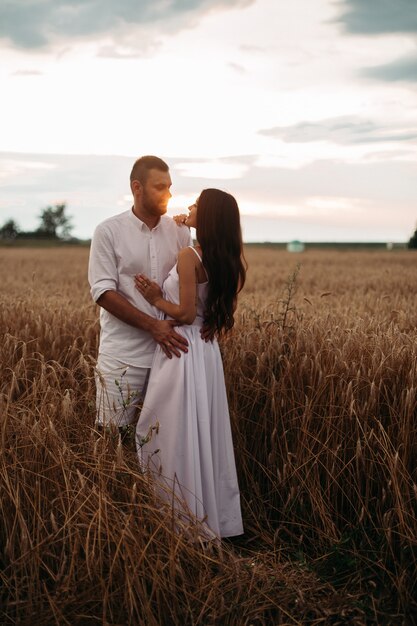 Stock photo pleine longueur d'un couple romantique en vêtements blancs étreignant dans le champ de blé au coucher du soleil.