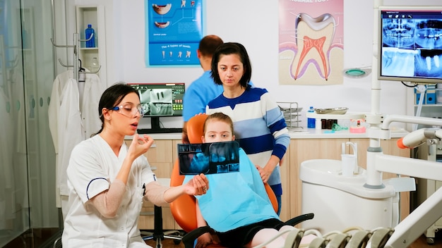 Stomatologue expliquant le traitement dentaire tenant une radiographie pointant sur les dents affectées pendant qu'un assistant prépare des outils stérilisés pour la chirurgie. Médecin et infirmière travaillant dans l'unité de stomatologie